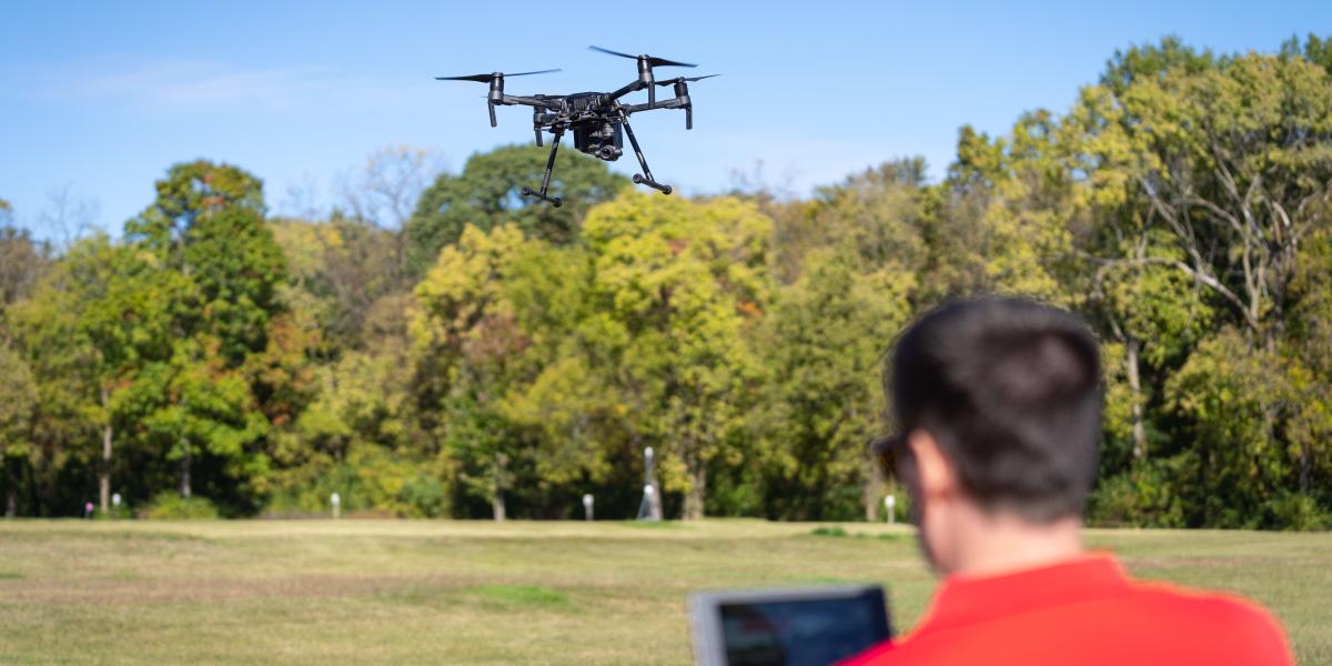 A man wearing a red shirt is seen out of focus, flying a large black drone.