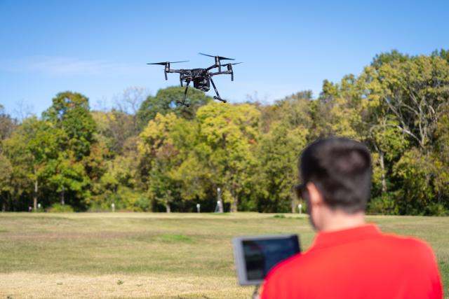 A man wearing a red shirt is seen out of focus, flying a large black drone.