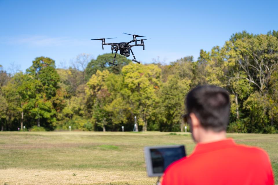 A man wearing a red shirt is seen out of focus, flying a large black drone.